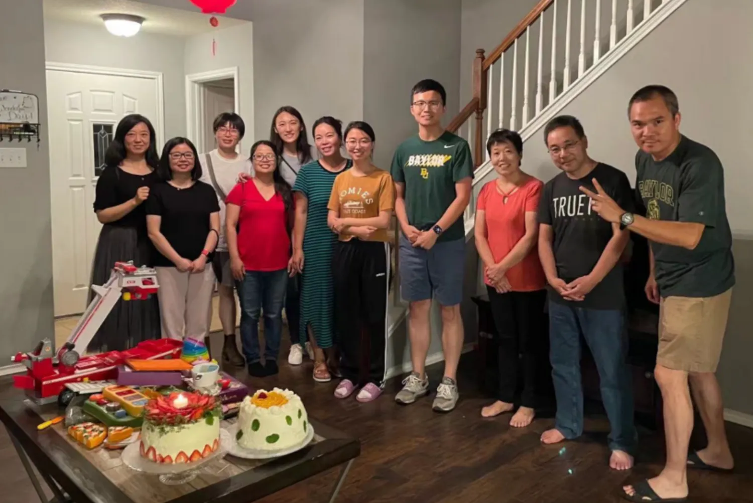约十二位弟兄姊妹站在客厅里合影，前方矮桌上放着两个装饰好的蛋糕，其中一个插着点燃的蜡烛。