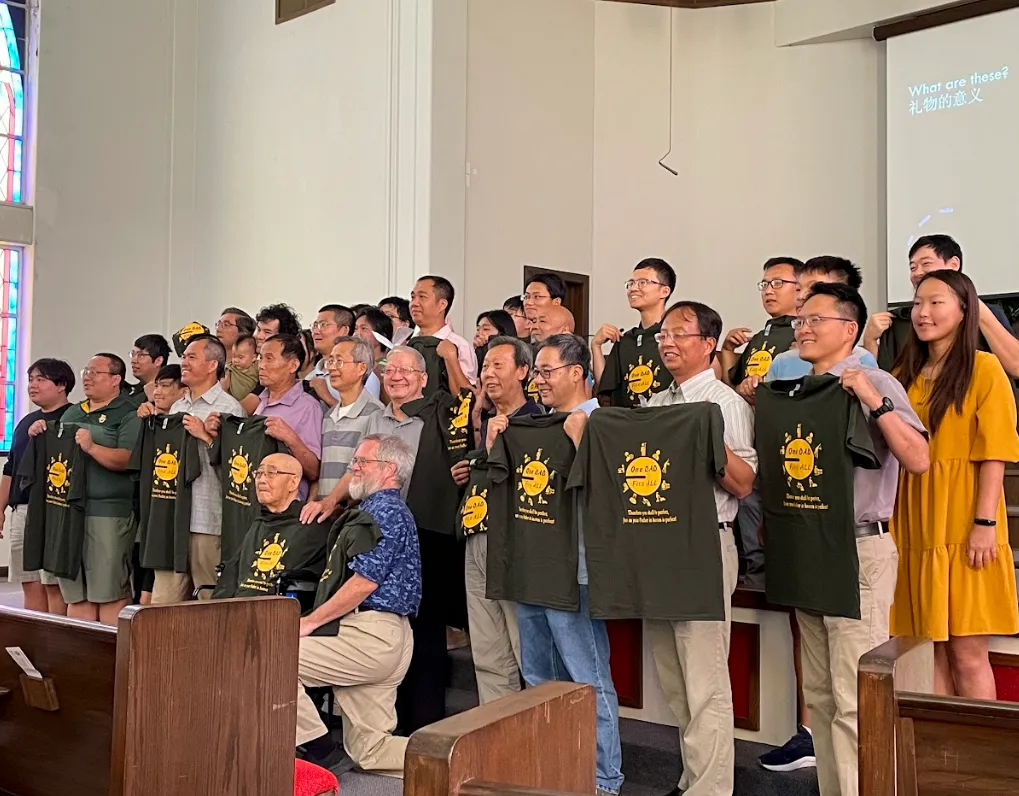 约二十五位弟兄，以及几位姊妹、一位少年和一个小孩站在礼堂里，举起印着 One DAD Fits ALL 的墨绿色 T 恤。