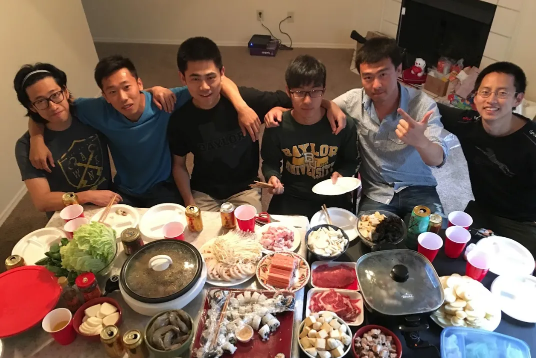 六位年轻弟兄在家中围坐矮桌旁，肩并着肩，桌上摆着两个电火锅、牛肉片、虾、豆腐和青菜。