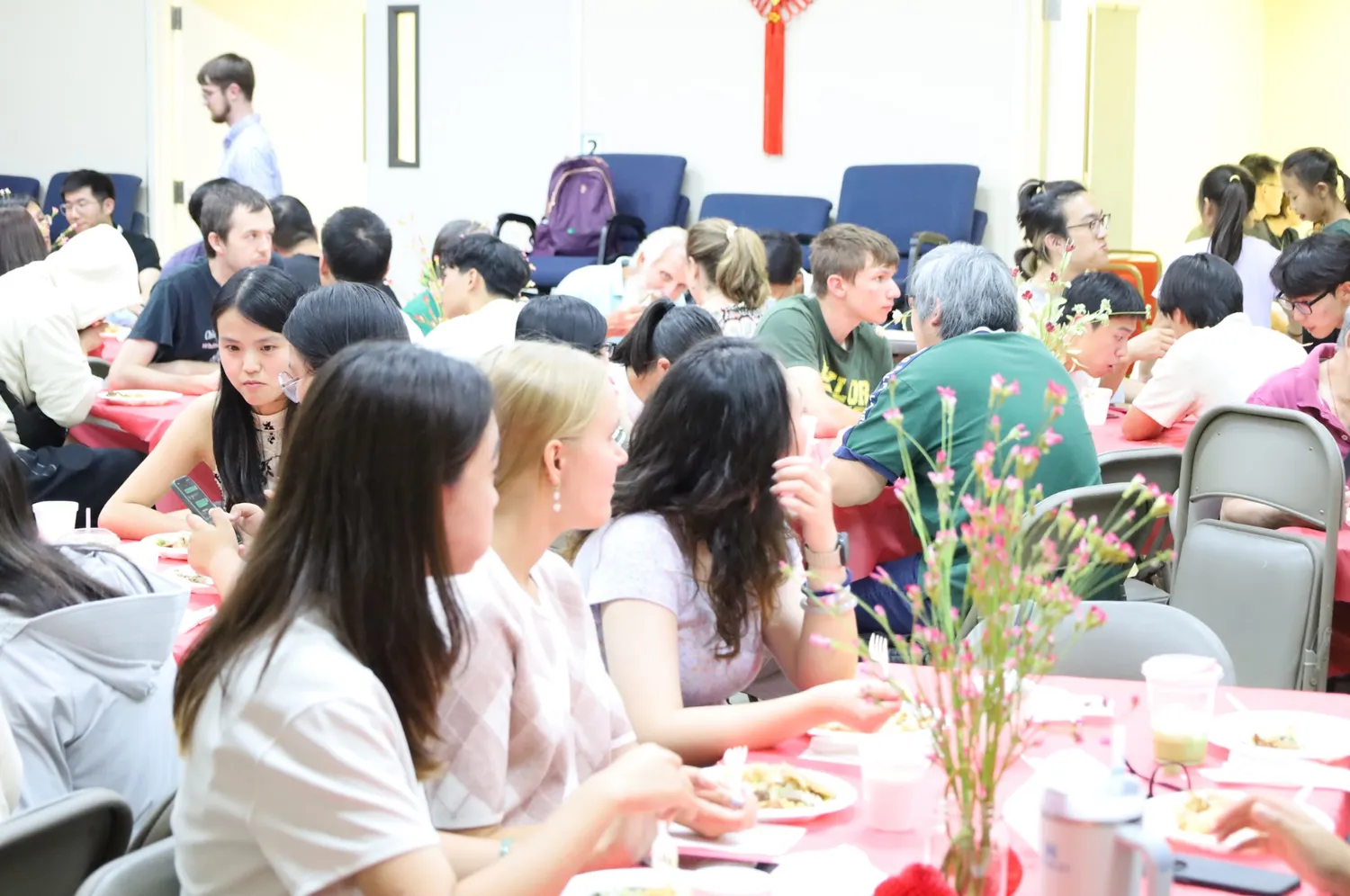 团契厅里五十多人围坐在铺着红桌布的长桌旁一同用餐，身后墙上挂着红色中国结。