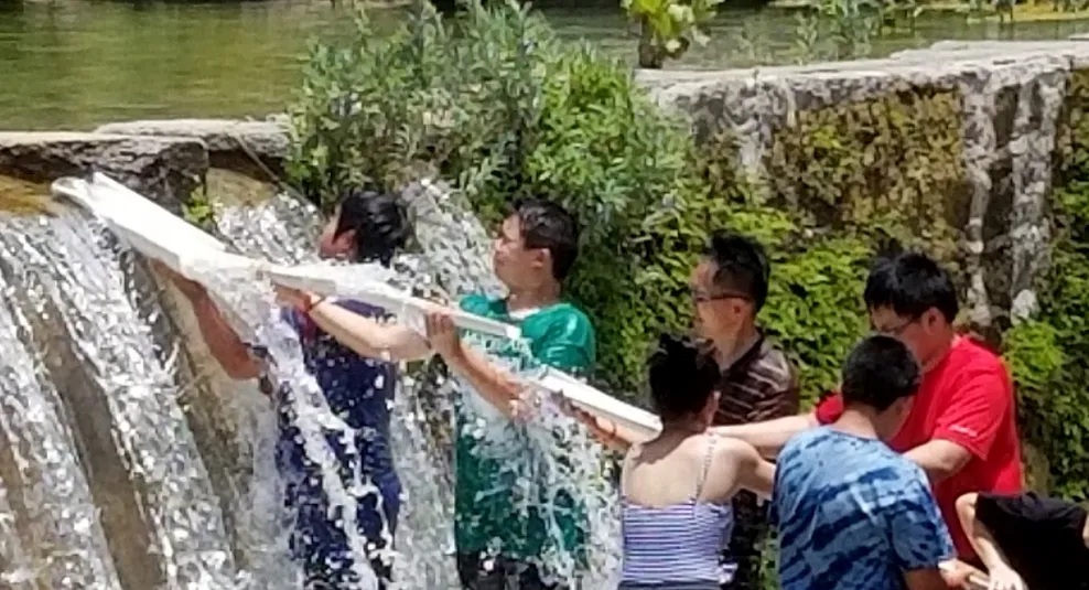 七个人站在瀑布下的浅溪中，合力传递一条长长的水槽，完成团队协作的活动。