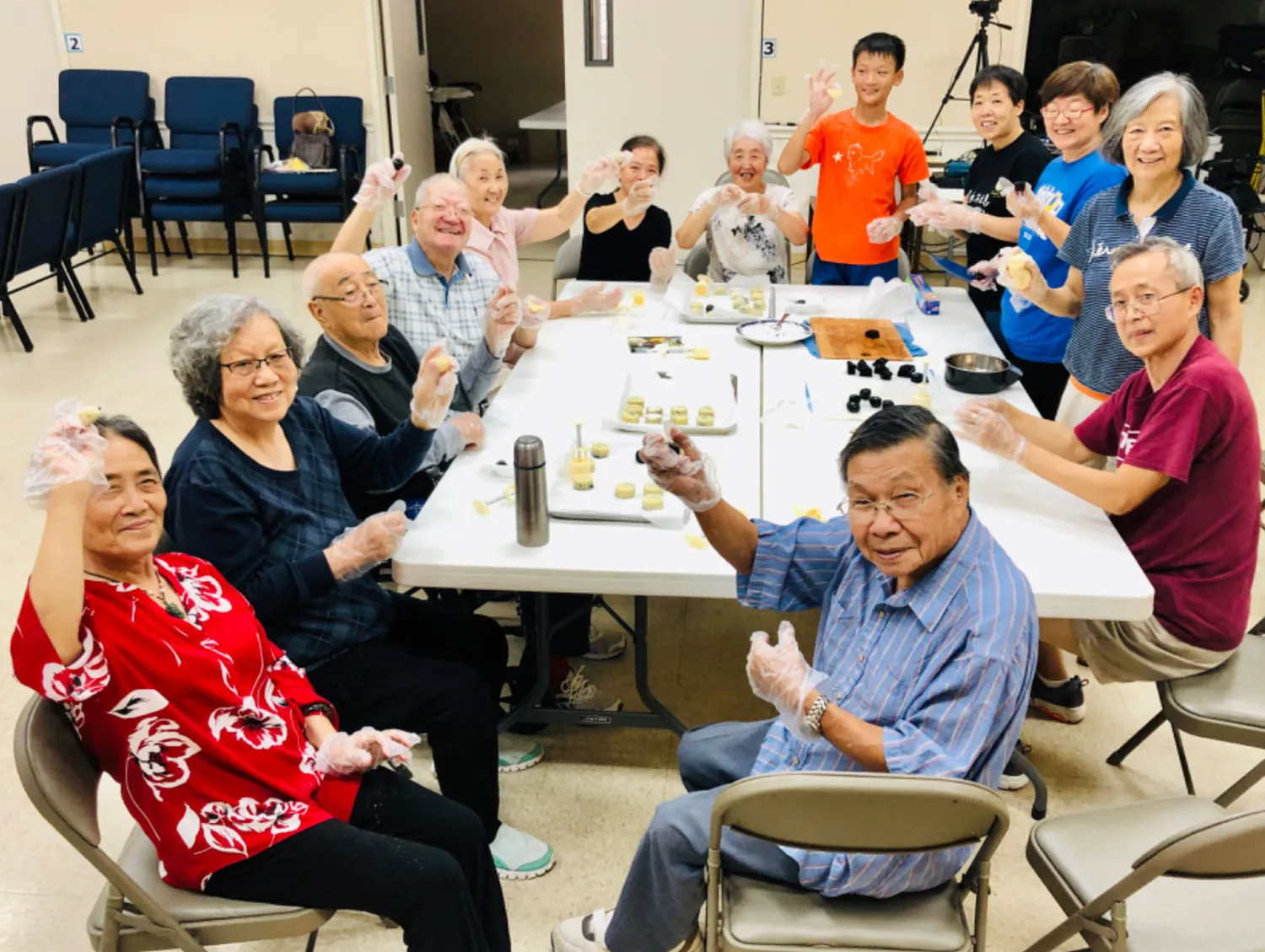 十几位年长的弟兄姊妹与一位男孩围在团契厅的折叠桌旁，戴着食品手套，把月饼压进模具里。